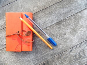 pencil and blue ink pen laid on an orange leather bound notebook with brown leather tie with gold leaf tassel laid on a wooden table