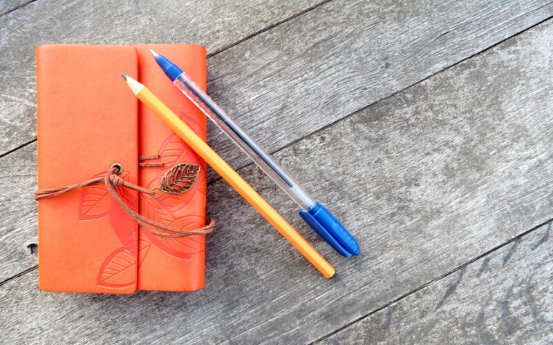 pencil and blue ink pen laid on an orange leather bound notebook with brown leather tie with gold leaf tassel laid on a wooden table