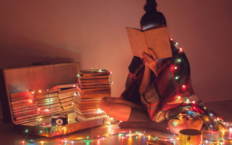 Young woman with her face in a book, sitting on the floor surrounded by books and a strand of Christmas lights