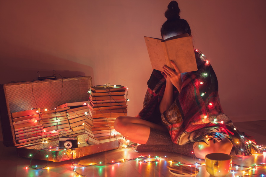 Young woman with her face in a book, sitting on the floor surrounded by books and a strand of Christmas lights