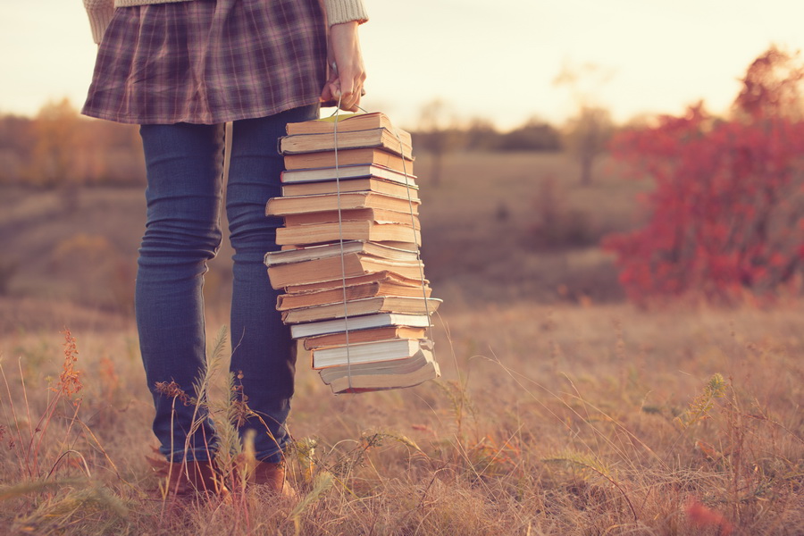 Woman standing in a field, carrying a stack of books bound in rope like a suitcase